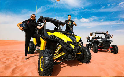 Two-seater desert safari buggy speeding across the vast, sun-soaked sand dunes, with both passengers wearing helmets and gripping the handlebars, creating a plume of sand in its wake against the picturesque desert landscape