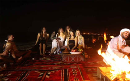 Under a star-studded desert sky, an overnight desert safari camp comes to life. Tents are illuminated with soft light, and a bonfire glows warmly in the center. Tourists gather around the fire, enjoying the night in the tranquil desert, with sand dunes stretching into the distance and the Milky Way shining brightly above.